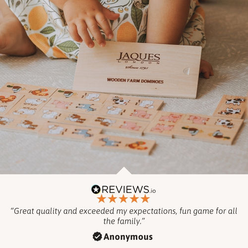 A child's hands and legs are visible as they play with Jaques London Wooden Farm Dominoes on a light-colored carpet. The wooden game pieces are spread out, and the wooden box with the brand name is held up. 