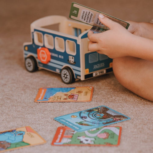 A child's hands holding a school bus card in a colorful educational game, with several illustrated cards scattered on a beige carpet next to a blue and cream toy school bus. 