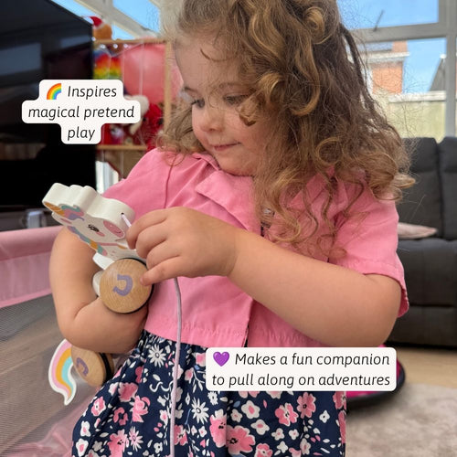 A young girl with curly light brown hair wearing a pink shirt and a navy floral skirt is shown in a living room setting, holding and playing with a wooden toy that has a rainbow-colored pull tab. 