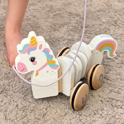 A wooden pull toy unicorn with a colorful painted face, mane, and rainbow tail, being pulled by a child's foot on a beige shag rug. 