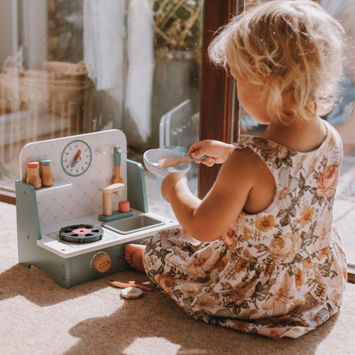 A young child with blonde hair sits on a carpeted floor by a window, playing with a wooden toy kitchen set. The child is wearing a sleeveless floral dress.