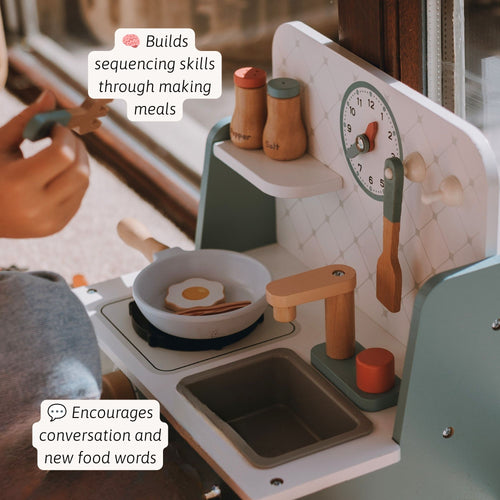 A child's wooden play kitchen with a white and sage green body, featuring a stovetop with a frying pan, a sink, shelves, and toy food items. A hand is visible holding a toy utensil near the kitchen, with text overlays highlighting developmental benefits.