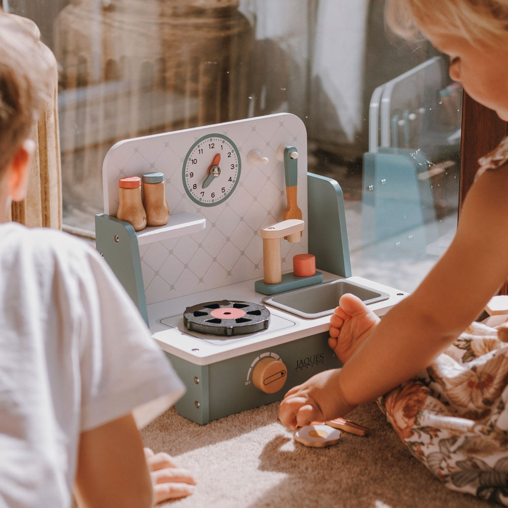A children's wooden play kitchen set with a mint green and white design, featuring a stove, sink, clock, and utensils, shown in a lifestyle setting with children playing near a window.