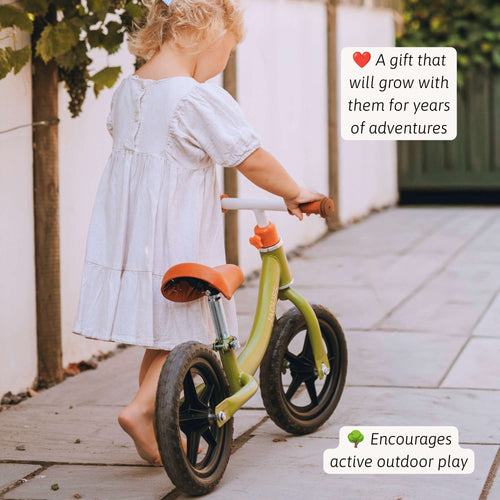 A young child in a white dress pushes a lime green balance bike with orange seat and handlebar along a paved outdoor path. Text overlays on the right describe the benefits of the bike. 