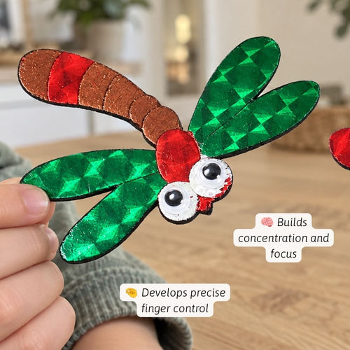 A close-up of a hand holding a handmade dragonfly craft. The dragonfly has green glitter wings, a brown glitter body, a red glitter head, and white googly eyes.