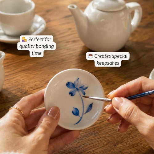 Person painting a blue flower on a white ceramic dish with teacup and teapot in the background.