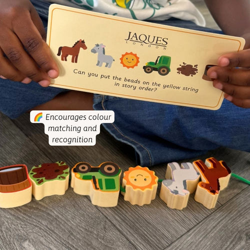 A person holds a printed instruction card for a wooden bead puzzle toy, with several wooden animal and vehicle beads arranged on a gray wood floor. The card shows the brand name JAQUES LONDON and a matching game question.