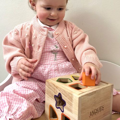 Child playing with a wooden shape sorting toy, wearing a pink outfit. 