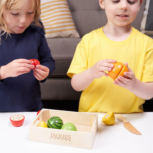 Two children playing with toy fruits and vegetables on a table, with a Jaques of London box in the foreground.  