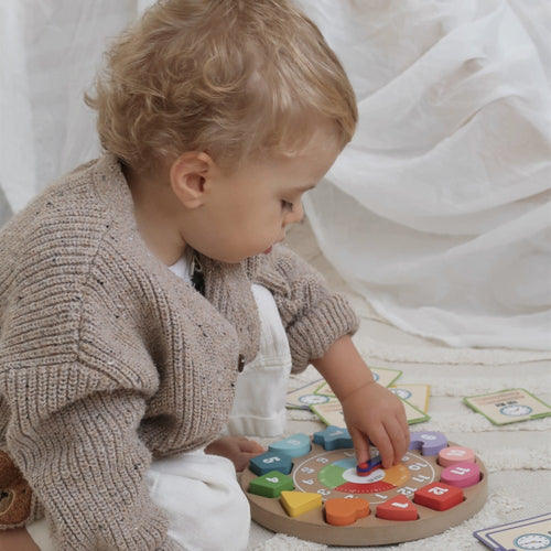 Young child fitting a coloured shape block into the Jaques of London wooden learning clock on a soft rug 